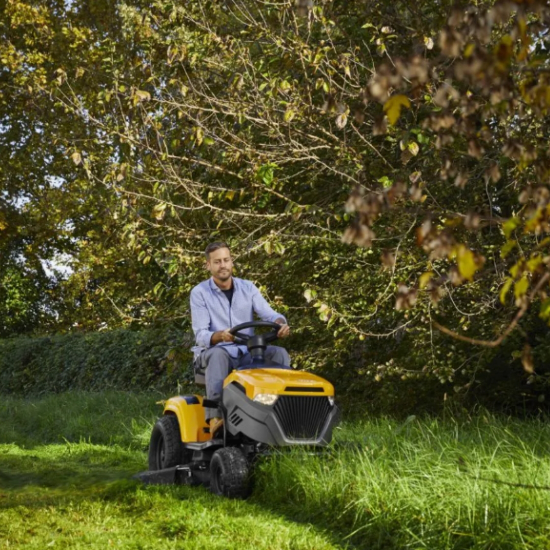Stiga Tornado 5108W petrol garden tractor mowing long grass in a large garden, driven by an operator through an open grassy area bordered by mature trees.