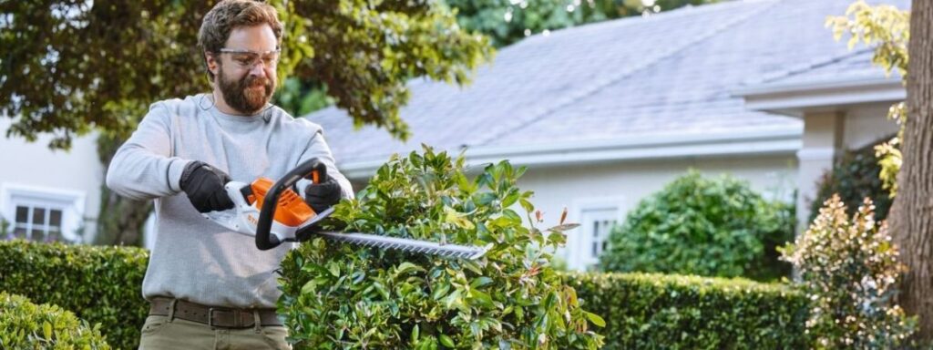 Person using a cordless hedge trimmer to shape a garden hedge, demonstrating the convenience and performance of battery-powered garden machinery.