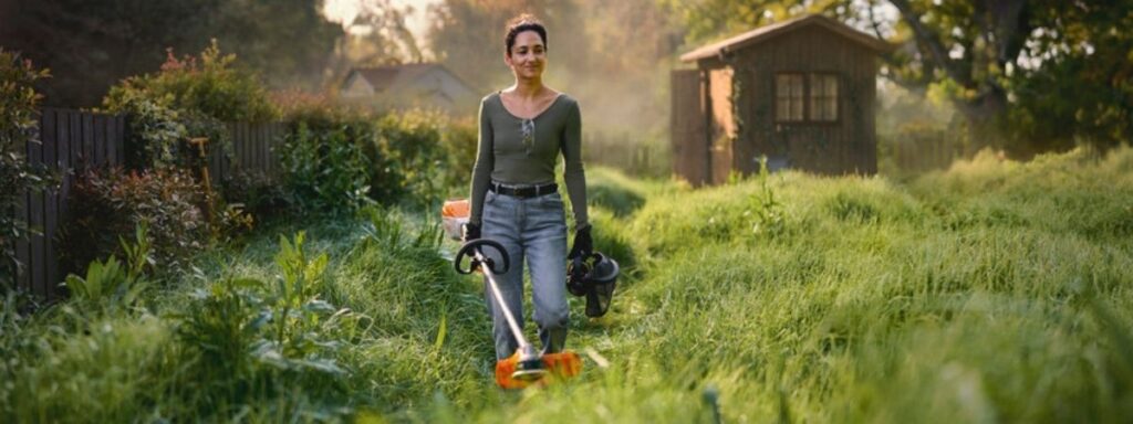 Battery-powered grass trimmer being used to cut long grass in a garden, showcasing modern cordless garden machinery.