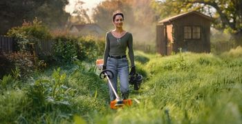 Woman using cordless garden tools in an overgrown garden with battery-powered grass trimmer and hedge trimmer.
