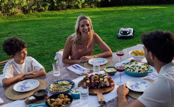 Family enjoying a meal in the garden while a robotic lawn mower maintains the lawn in the background.